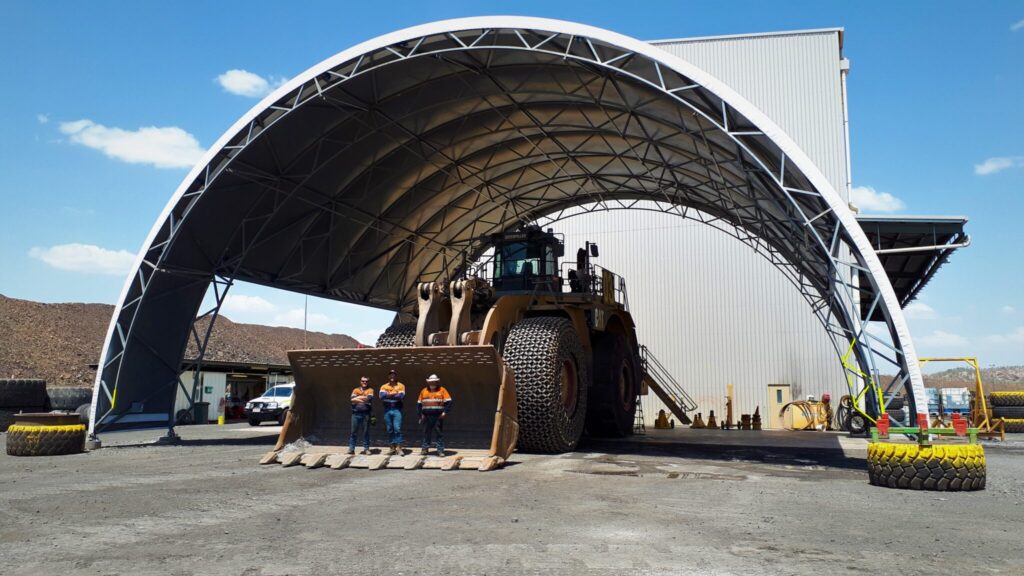 A wide fabric shelter supported by shipping containers, creating a shaded and ventilated workspace to protect workers and equipment from the intense summer heat.