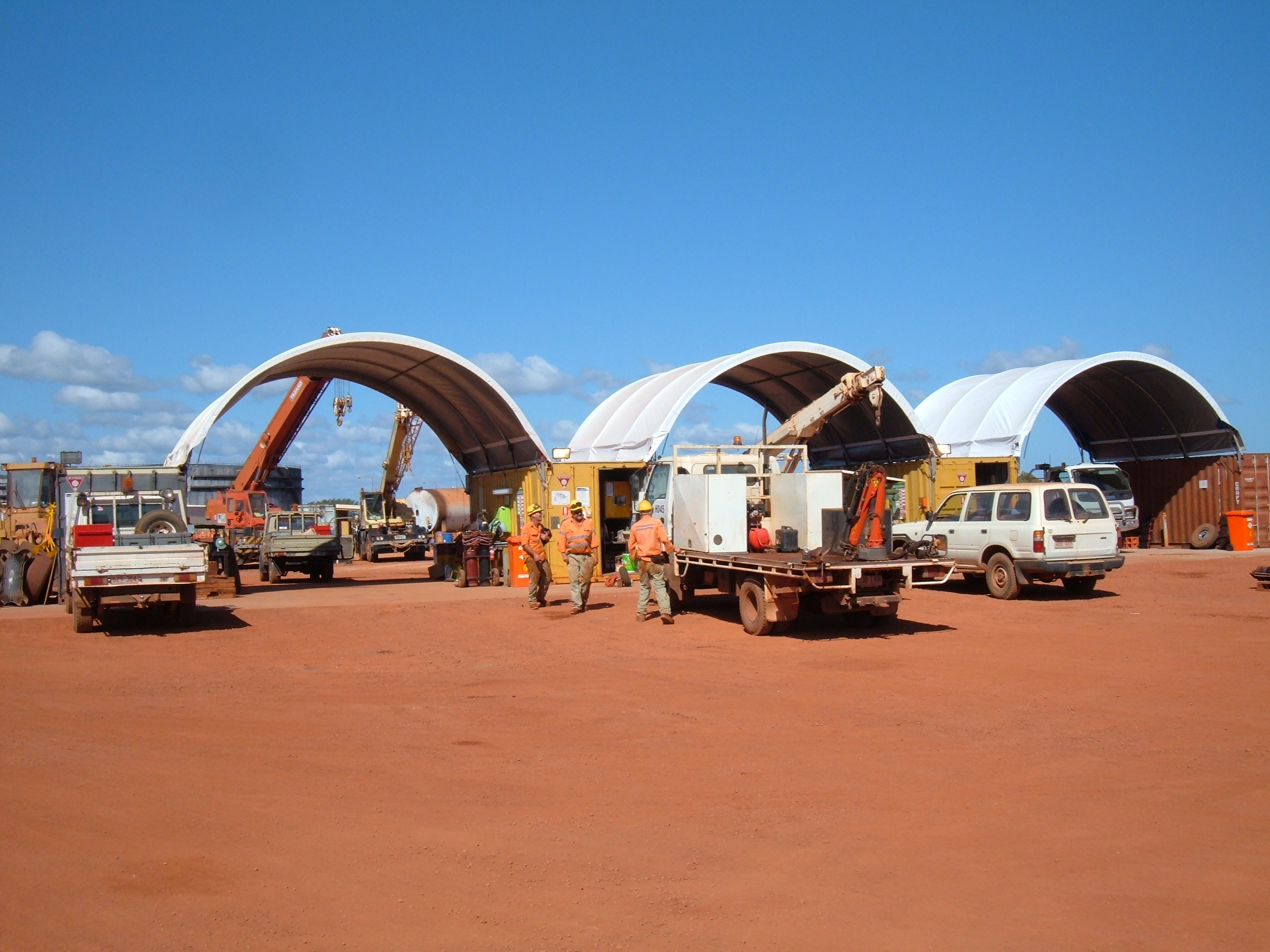 A series of container domes sheltering equipment and vehicles on a red soil construction site under clear skies.