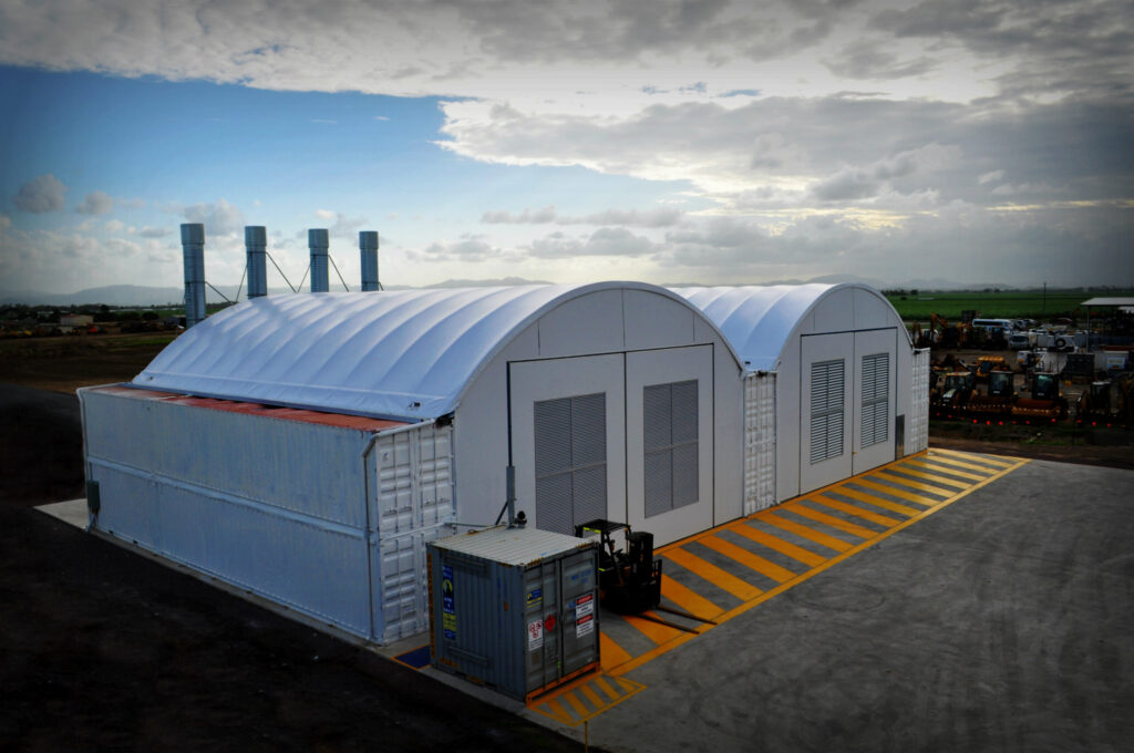 White double-arched fabric shelter built on shipping containers at an industrial site, with ventilation panels and safety markings.