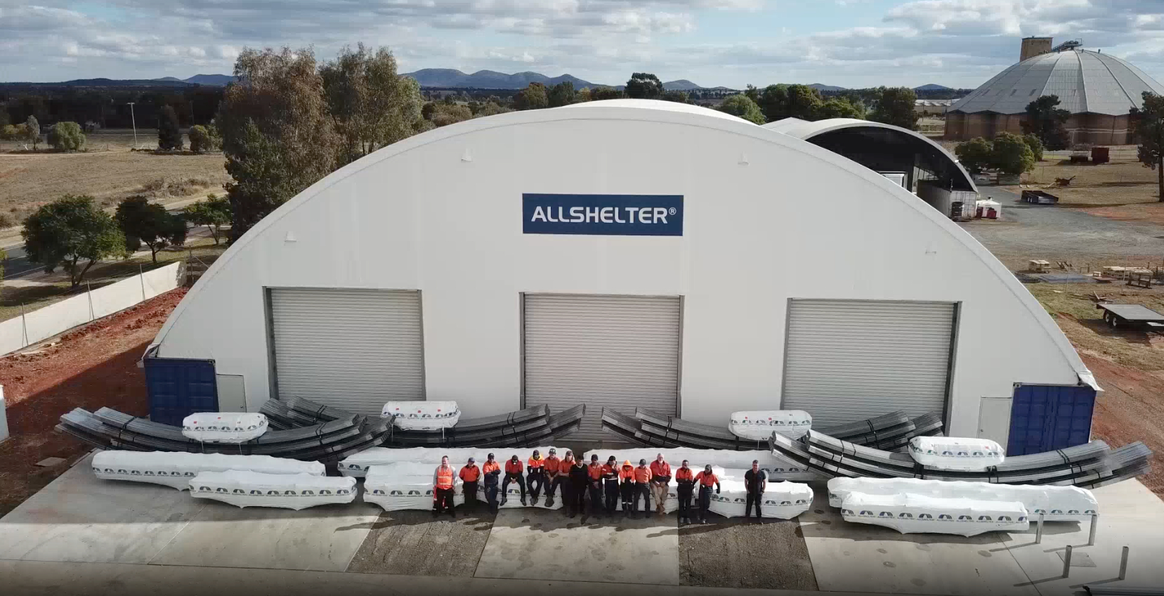 A large Allshelter fabric shelter with a team of workers and shelter components displayed in front.